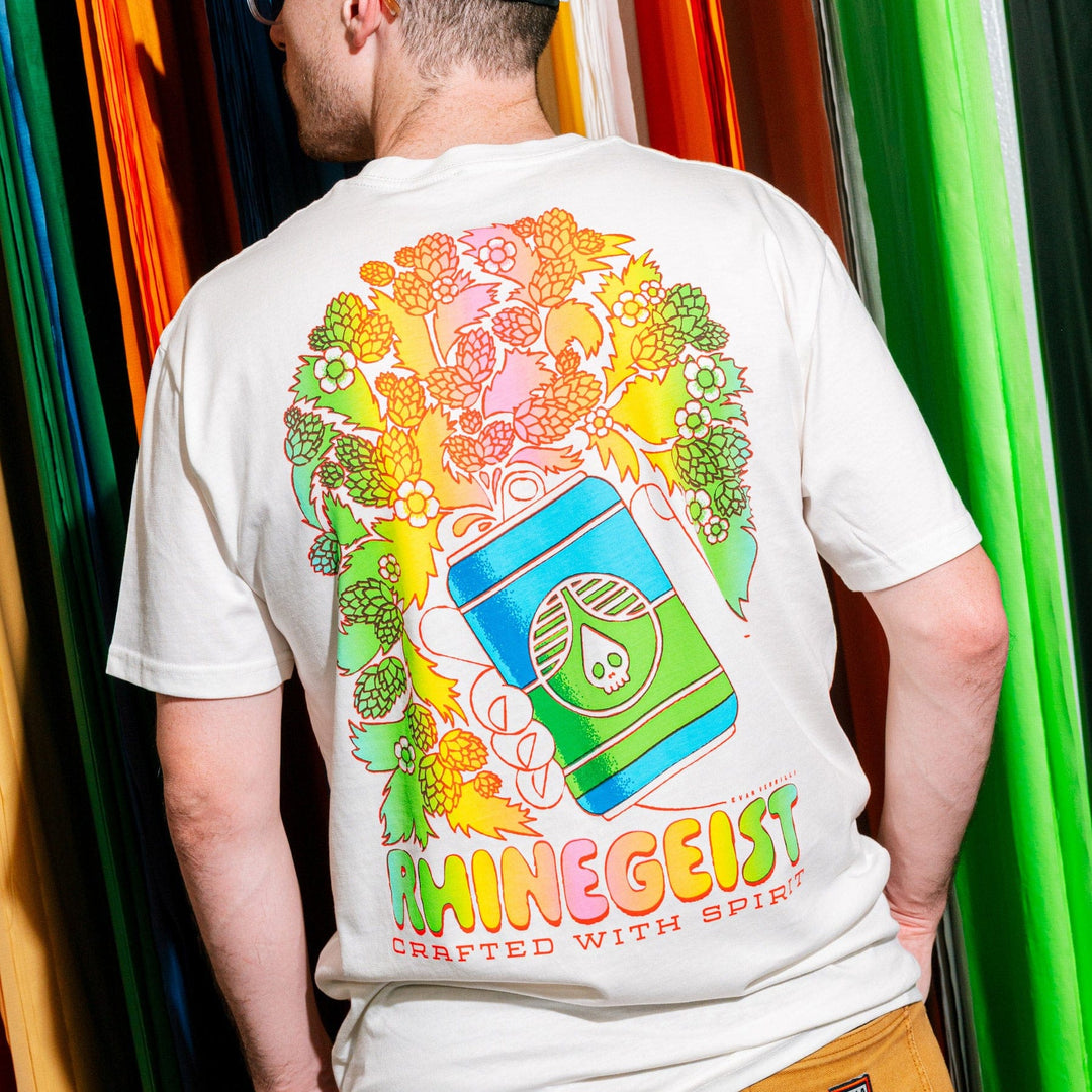 Person wearing a white t-shirt with colorful graphics and 'Rhinegeist' branding in front of colorful curtains.