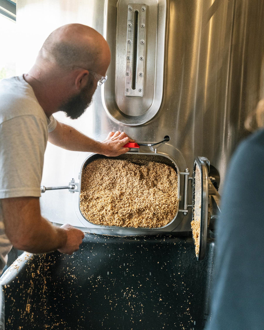 Person working with grain in a large metal container, likely in a brewery setting.